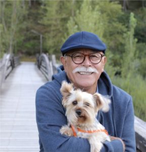 Apartments in Lebanon A smiling man wearing glasses and a blue cap holding a small dog on a wooden bridge outdoors.