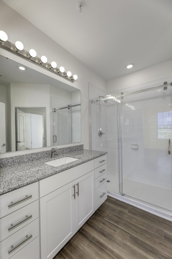 Apartments in Lebanon, IN Modern bathroom with a granite countertop vanity, large mirror with globe lights, white cabinets, and a glass-enclosed shower. The floor has wood-style tiles.