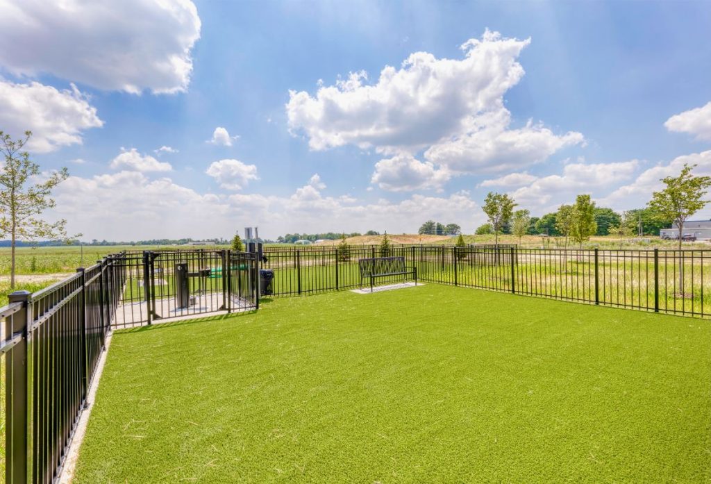 Apartments in Lebanon, IN A fenced outdoor area with artificial grass, surrounded by open fields, trees, and a clear blue sky with scattered clouds.