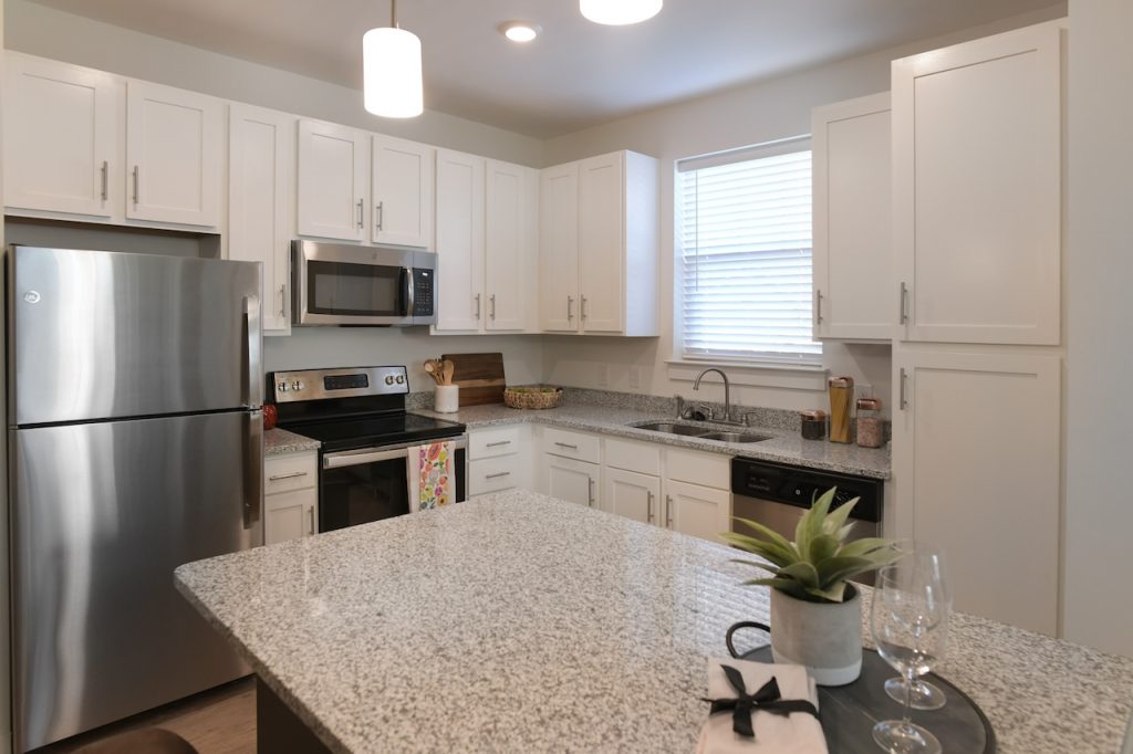 Apartments in Lebanon, IN Modern kitchen in brand new apartments in Lebanon, IN, featuring white cabinets, stainless steel appliances, and granite countertops. An island with a small plant and wine glasses graces the foreground while blinds cover the window above the sink.