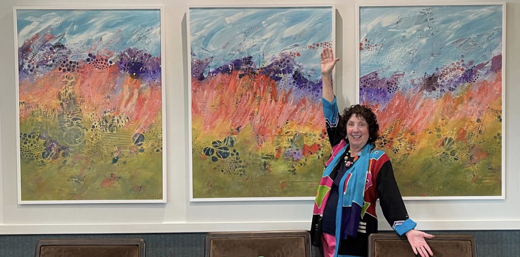 Apartments in Lebanon, IN A person stands smiling with one arm raised in front of a colorful three-panel abstract painting, like an Auto Draft masterpiece, mounted on a wall. The painting features vibrant splashes of reds, blues, and greens.