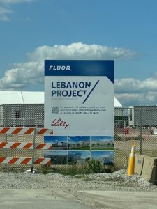 Apartments in Lebanon, IN A construction site sign reads "FLUOR Lebanon Project" with contact information and logos for Fluor and Lilly. Highlighting optimal living for Eli Lilly's biotech professionals, the sign features images of construction and is placed next to a fenced area with orange barriers.