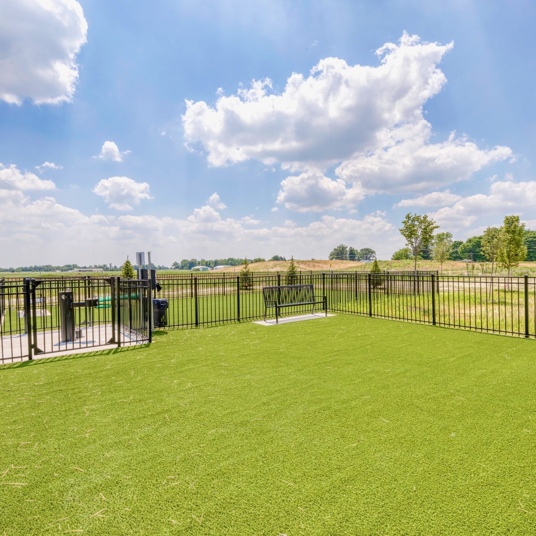 Apartments in Lebanon, IN Outdoor area with a fenced-in artificial grass space under a partly cloudy sky, surrounded by greenery and distant trees—a perfect feature to highlight in any realtor's listing.