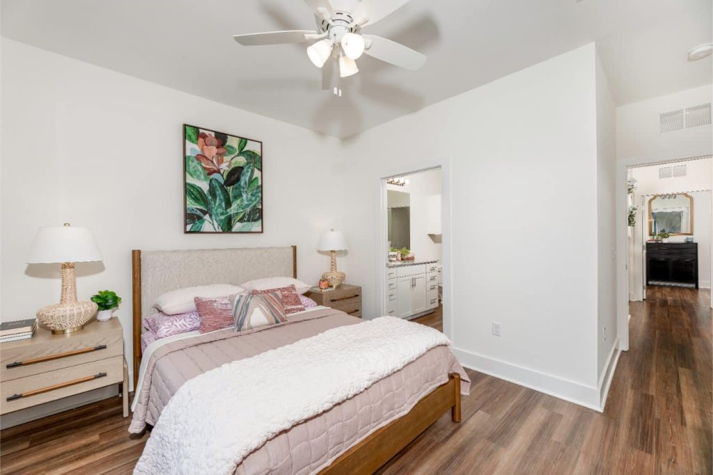 Apartments in Lebanon, IN A neatly arranged bedroom with a double bed, bedside tables, lamps, a ceiling fan, and a colorful painting above the bed. The room has wooden flooring and an open view into an adjacent bathroom.