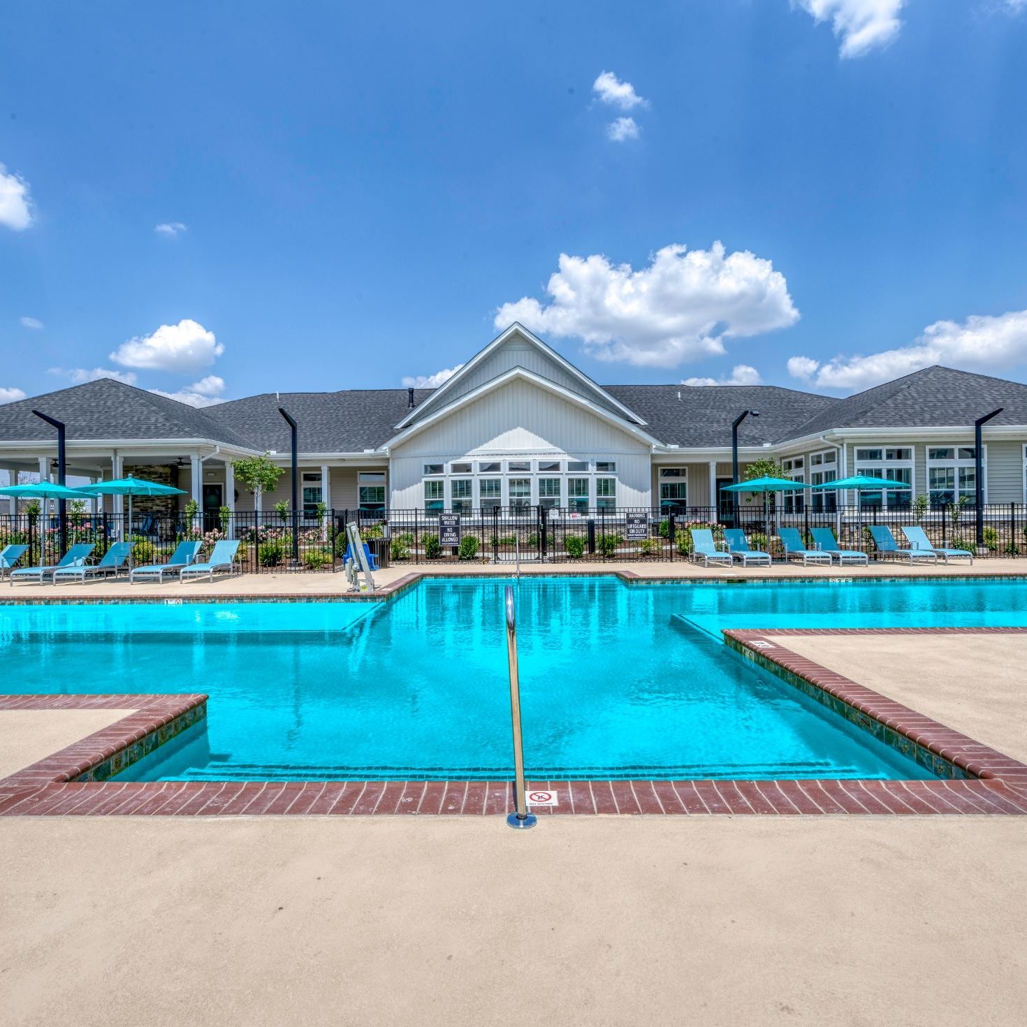 Apartments in Lebanon, IN Outdoor swimming pool with lounge chairs and umbrellas surrounding it, in front of a large community building under a blue sky with scattered clouds, creating a perfect home-like atmosphere.