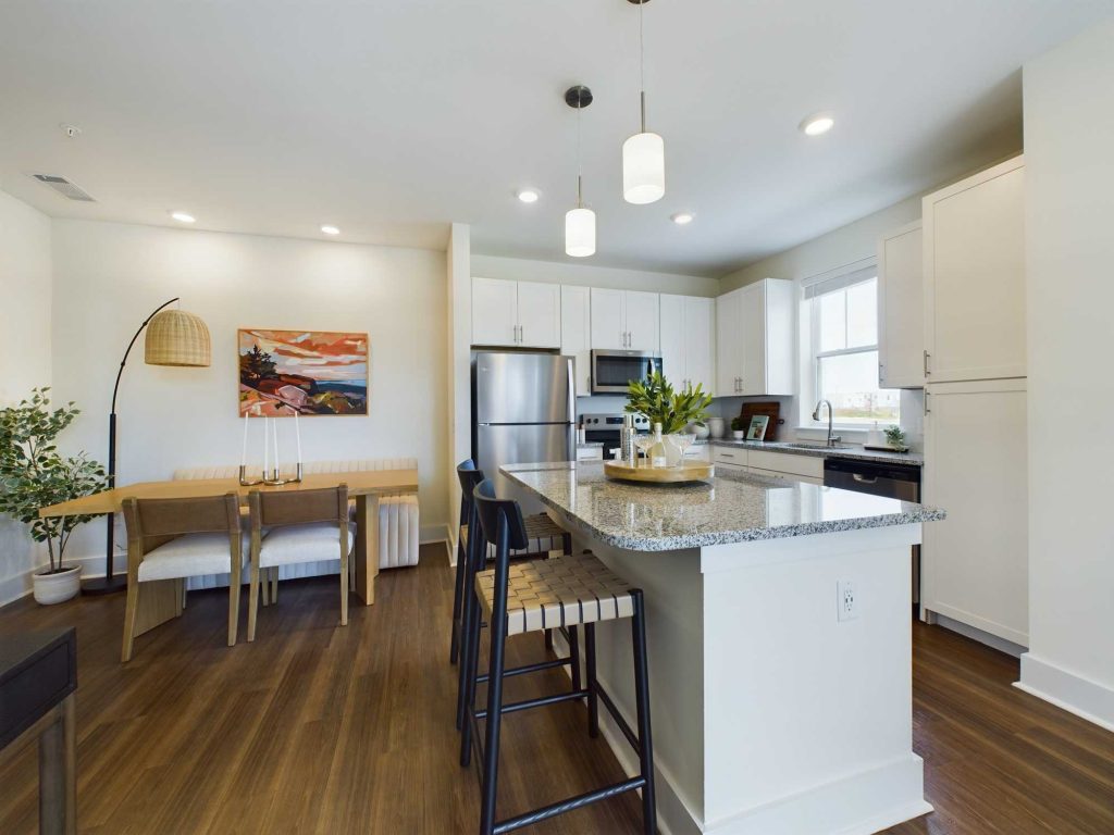 Apartments in Lebanon, IN Modern kitchen and dining area featuring white cabinets, stainless steel appliances, a granite island with bar seating, wooden dining table with chairs, and pendant lighting. Artwork and plants adorn the space.