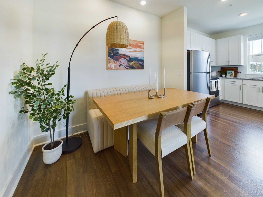 Apartments in Lebanon, IN A modern dining area with a wooden table, four chairs, a potted plant, and a hanging lamp. The room features white walls and a kitchen with stainless steel appliances in the background.