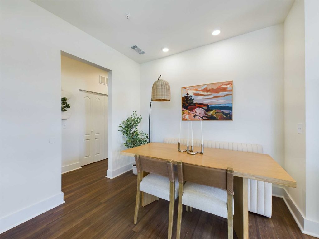 Apartments in Lebanon, IN A small dining area with a wooden table, three chairs, a bench, a potted plant, a floor lamp, and a colorful landscape painting on a white wall. A doorway leads to another room.