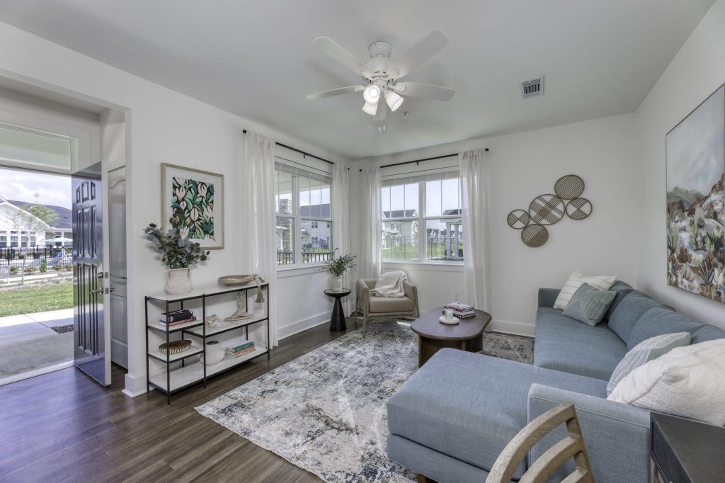 Apartments in Lebanon, IN A bright living room in this two-bedroom apartment features a blue sectional sofa, a round wooden coffee table, a patterned rug, a bookshelf, and wall art. Large windows allow natural light to flood in, and a ceiling fan hangs overhead.