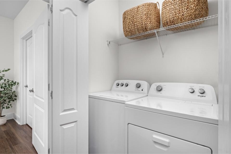 Indiana, Boone County, Lebanon Apartments for rent A laundry room with a white washer and dryer, a wire shelf holding two woven baskets above, white walls, and a plant in the corner. See all available apartments for rent at Hickory Cottages in Indiana Boone County Lebanon, Hickory Cottages has rental units ranging from 664-1420 sq ft