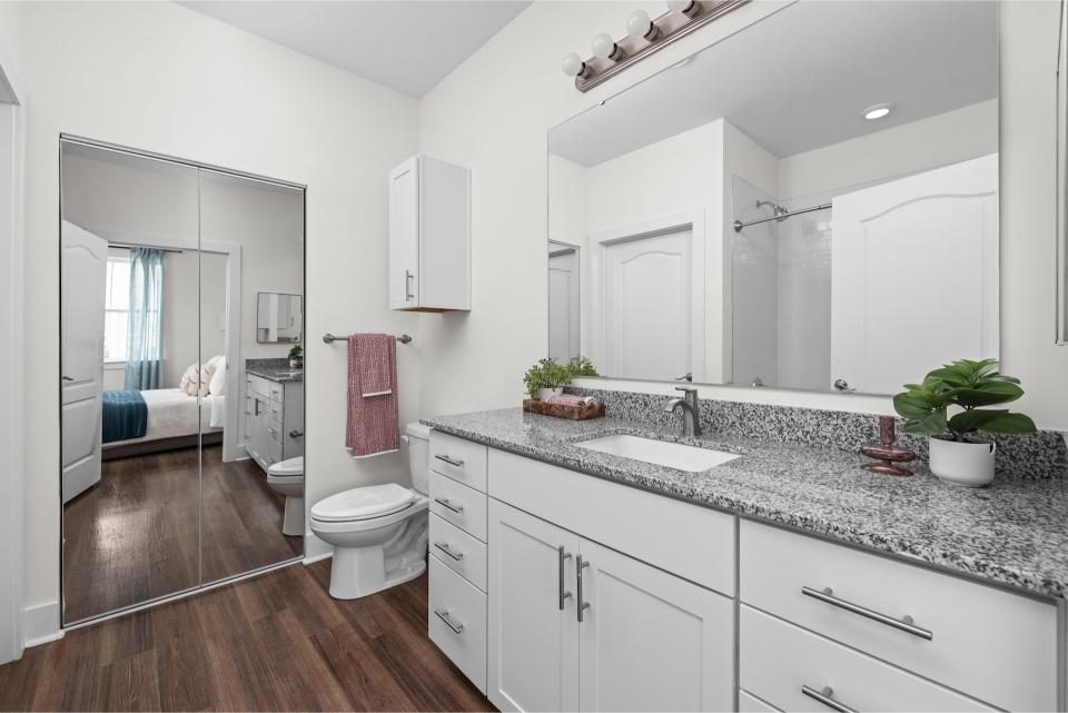 Indiana, Boone County, Lebanon Apartments for rent Modern bathroom with white cabinets, granite countertop, a large mirror, wooden floor, and a view into a bedroom through a mirrored sliding closet door. See all available apartments for rent at Hickory Cottages in Indiana Boone County Lebanon, Hickory Cottages has rental units ranging from 664-1420 sq ft