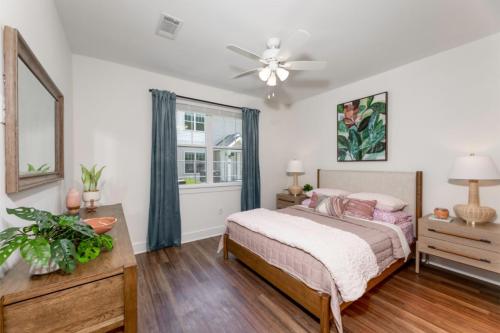 Apartments in Lebanon, IN A bedroom with a bed, two bedside tables with lamps, a dresser with plants, a ceiling fan, a window with blue curtains, and a leafy wall art. The room has wooden flooring and white walls.