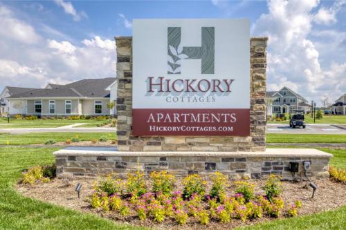 Apartments in Lebanon, IN A sign for Hickory Cottages Apartments with a landscaped base, standing in front of apartment buildings on a sunny day. The website hickorycottages.com is visible at the bottom of the sign.