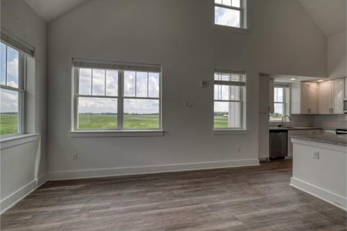 Apartments in Lebanon, IN A bright, empty room with large windows overlooking a grassy field, featuring hardwood floors and an adjacent modern kitchen with white cabinets and stainless-steel appliances.