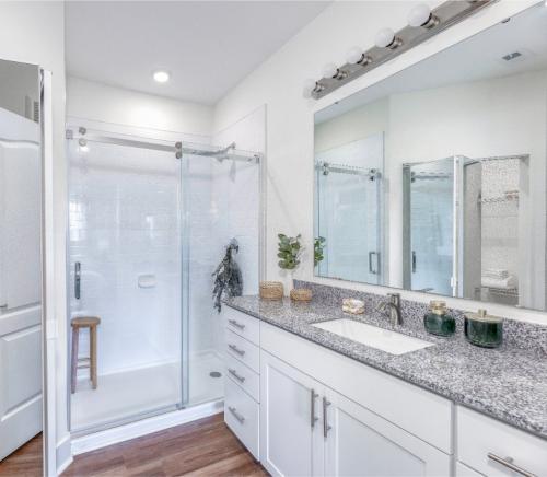 Apartments in Lebanon, IN A bathroom with a walk-in glass shower, a granite countertop with a sink, storage cabinets, a mirror with lights above it, and a wooden stool inside the shower. White walls and wood flooring.