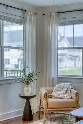 Apartments in Lebanon, IN A cozy corner with a tan armchair, a knitted throw, a small round table with a potted plant, and sheer curtains framing two large windows. Sunlight brightens the room.