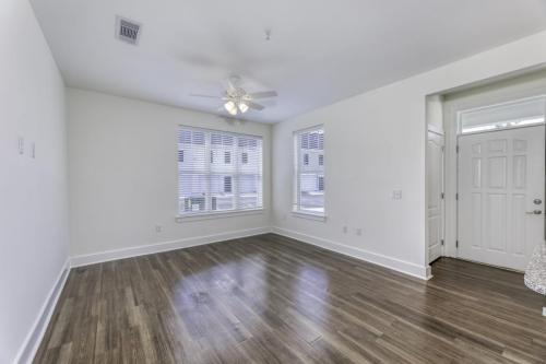 Apartments in Lebanon, IN A vacant room with light-colored walls, wood flooring, two large windows with blinds, a ceiling fan, and a white front door with a window panel.