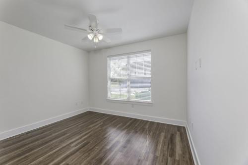 Apartments in Lebanon, IN An empty room with wood flooring, a ceiling fan with lights, white walls, and a large window with blinds.