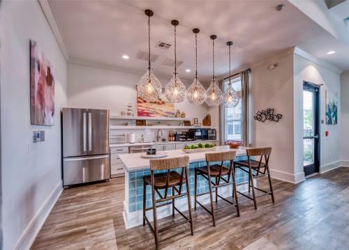 Apartments in Lebanon, IN Modern kitchen with an island, four wooden barstools, stainless steel refrigerator, pendant lights, and wall art. The flooring is light wood and the space has large windows bringing in natural light.