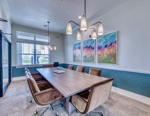 Apartments in Lebanon, IN A conference room with a wooden table, six brown chairs, a modern light fixture, large windows with blinds, and three colorful abstract paintings on the wall.