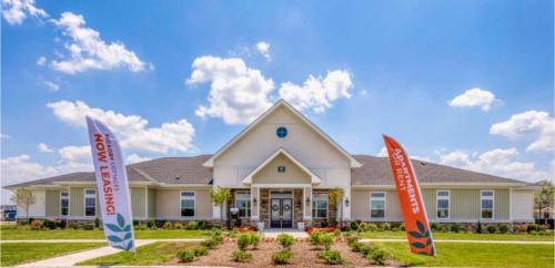 Apartments in Lebanon, IN A modern, single-story building with a gray roof, white siding, and a landscaped front yard. Two flags on either side of a pathway advertise leasing availability.