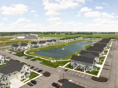 Indiana, Boone County, Lebanon Apartments for rent Aerial view of a suburban neighborhood with rows of houses surrounding a central pond with fountains, parking lots, and green fields in the background under a partly cloudy sky. See all available apartments for rent at Hickory Cottages in Indiana Boone County Lebanon, Hickory Cottages has rental units ranging from 664-1420 sq ft