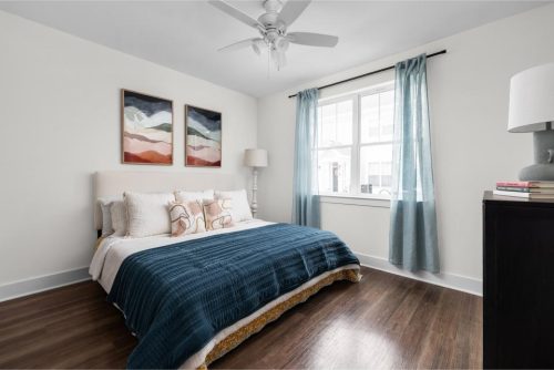 Indiana, Boone County, Lebanon Apartments for rent A bright bedroom with a bed covered in a blue blanket, decorative pillows, two framed artworks above the headboard, a window with blue curtains, and a ceiling fan. See all available apartments for rent at Hickory Cottages in Indiana Boone County Lebanon, Hickory Cottages has rental units ranging from 664-1420 sq ft