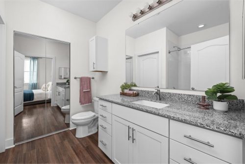 Indiana, Boone County, Lebanon Apartments for rent Modern bathroom with white cabinets, granite countertop, a large mirror, wooden floor, and a view into a bedroom through a mirrored sliding closet door. See all available apartments for rent at Hickory Cottages in Indiana Boone County Lebanon, Hickory Cottages has rental units ranging from 664-1420 sq ft
