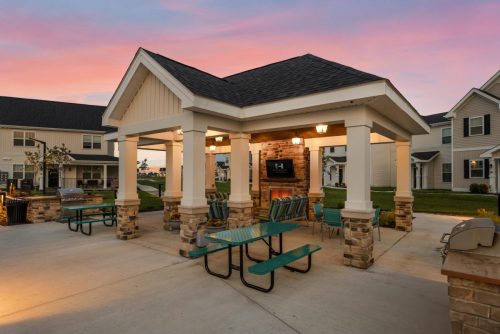 Indiana, Boone County, Lebanon Apartments for rent Covered outdoor pavilion with picnic tables, chairs, and a wall-mounted TV, surrounded by multi-story residential buildings at sunset. See all available apartments for rent at Hickory Cottages in Indiana Boone County Lebanon, Hickory Cottages has rental units ranging from 664-1420 sq ft