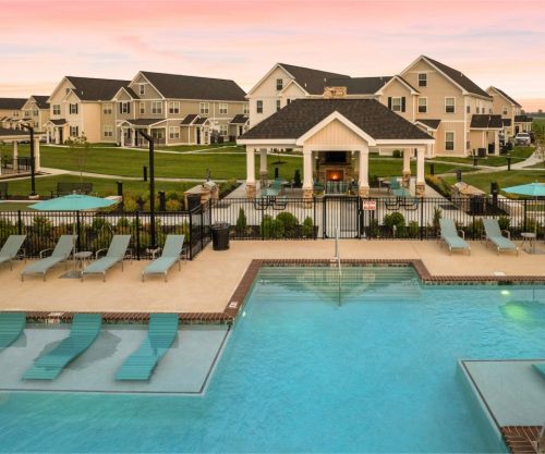Indiana, Boone County, Lebanon Apartments for rent Outdoor swimming pool area with lounge chairs, surrounded by a fence, with a clubhouse and residential buildings in the background under a pink sunset sky. See all available apartments for rent at Hickory Cottages in Indiana Boone County Lebanon, Hickory Cottages has rental units ranging from 664-1420 sq ft