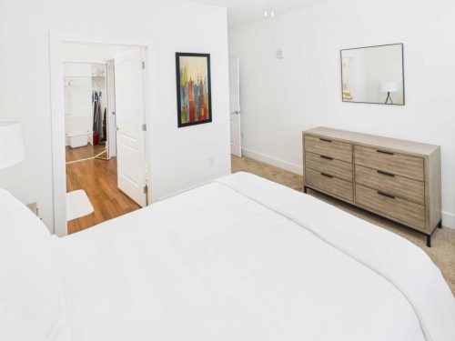 Indiana, Boone County, Lebanon Apartments for rent A bright, minimalist bedroom with a white bed, wooden dresser, wall art, and an open door leading to a walk-in closet with wooden flooring. See all available apartments for rent at Hickory Cottages in Indiana Boone County Lebanon, Hickory Cottages has rental units ranging from 664-1420 sq ft