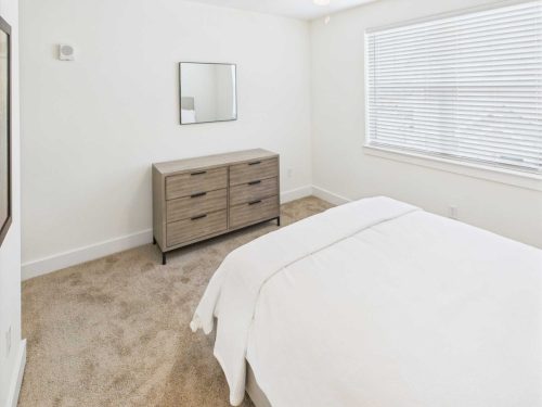 Indiana, Boone County, Lebanon Apartments for rent A minimalist bedroom with a bed covered in white bedding, a wooden dresser with a mirror above it, beige carpet, and a large window with closed blinds. See all available apartments for rent at Hickory Cottages in Indiana Boone County Lebanon, Hickory Cottages has rental units ranging from 664-1420 sq ft