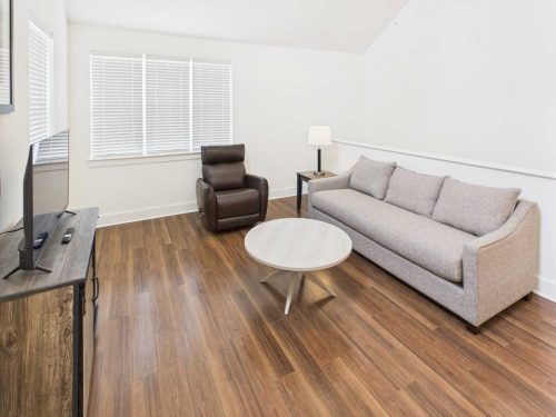 Indiana, Boone County, Lebanon Apartments for rent A modern living room with wood flooring, a gray sofa, a brown recliner, a round coffee table, a TV on a console, and a lamp on a side table near the window with closed blinds. See all available apartments for rent at Hickory Cottages in Indiana Boone County Lebanon, Hickory Cottages has rental units ranging from 664-1420 sq ft