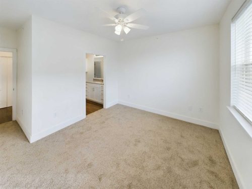 Indiana, Boone County, Lebanon Apartments for rent Empty bedroom with beige carpet, white walls, a ceiling fan with lights, large window, and a doorway leading to a bathroom with a visible vanity and mirror. See all available apartments for rent at Hickory Cottages in Indiana Boone County Lebanon, Hickory Cottages has rental units ranging from 664-1420 sq ft