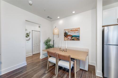 Apartments in Lebanon, IN A modern dining area featuring a wooden table with four chairs, a wall painting, a hanging light fixture, and a plant in the corner. The area is next to a stainless steel refrigerator.