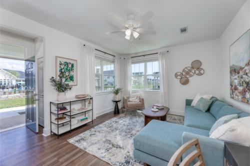 Apartments in Lebanon, IN Bright living room with hardwood floors, a blue sectional sofa, shelves with decor, armchair, wall art, patterned rug, and ceiling fan. Large windows offer views of greenery and outdoor space.