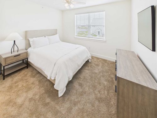 Indiana, Boone County, Lebanon Apartments for rent A bedroom with a large bed, white bedding, a wooden nightstand with a lamp, a dresser, a ceiling fan, and a window with blinds. See all available apartments for rent at Hickory Cottages in Indiana Boone County Lebanon, Hickory Cottages has rental units ranging from 664-1420 sq ft