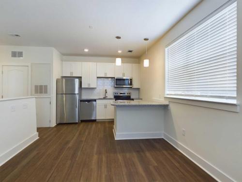 Apartments in Lebanon, IN Modern kitchen with white cabinets, stainless steel appliances, a gray countertop, and hardwood flooring. A large window with blinds is on the right side, and pendant lights hang above the countertop.