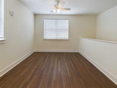 Apartments in Lebanon, IN An empty room with light-colored walls, a window with closed blinds, a ceiling fan with lights, and wooden flooring.