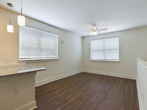 Apartments in Lebanon, IN Empty room with wood flooring, white walls, a ceiling fan, and two windows with blinds. Kitchen counter with hanging pendant lights visible on the left.