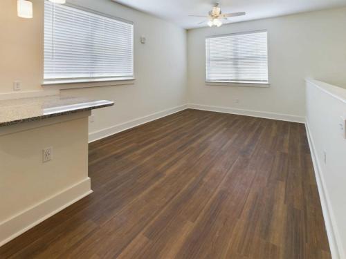 Apartments in Lebanon, IN A vacant room with light-colored walls, large windows with blinds, wooden flooring, a ceiling fan, and a kitchen counter visible on the left.