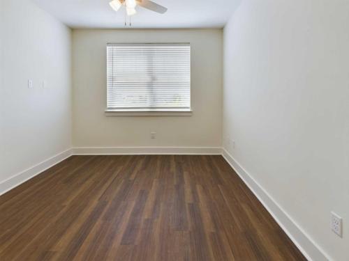 Apartments in Lebanon, IN An empty room with white walls, a ceiling fan, a small window with blinds, and dark wood flooring.