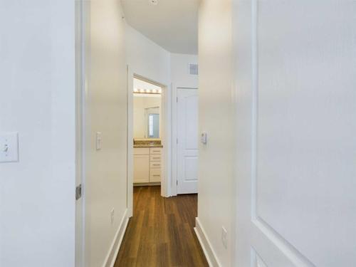 Apartments in Lebanon, IN A hallway with wooden flooring leads to a bathroom with a sink and mirror. The walls and doors are painted white. The bathroom vanity has a granite countertop.