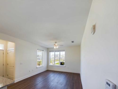 Apartments in Lebanon, IN Bright and airy room with hardwood flooring, white walls, a ceiling fan, two windows, and an open door leading to another area. A segment of a granite countertop is visible on the left.