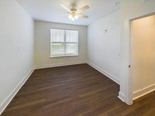 Apartments in Lebanon, IN A small, empty room with white walls, a ceiling fan, a window with blinds, and wooden flooring. An open door on the right leads to another area.