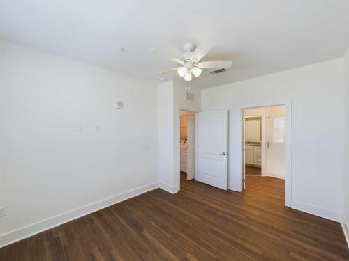 Apartments in Lebanon, IN An empty, white-walled room with a wooden floor, a ceiling fan with lights, and an open door leading to another room with a vanity and shower.