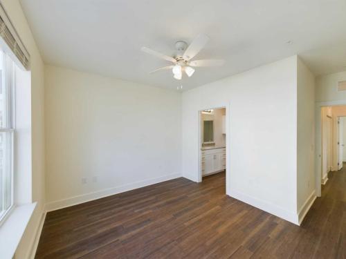 Apartments in Lebanon, IN An empty room with white walls, a ceiling fan with lights, a large window on the left, and an open doorway leading to a bathroom. The floor is wood, and another small hallway is visible on the right.