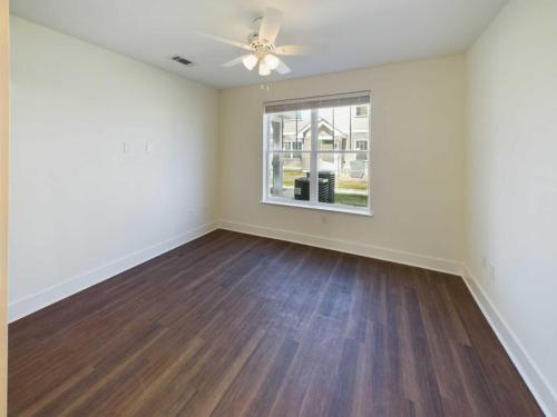 Apartments in Lebanon, IN Empty room with polished wooden floor, white walls, a ceiling fan with lights, and a large window showing a view of neighboring buildings and a grassy area.