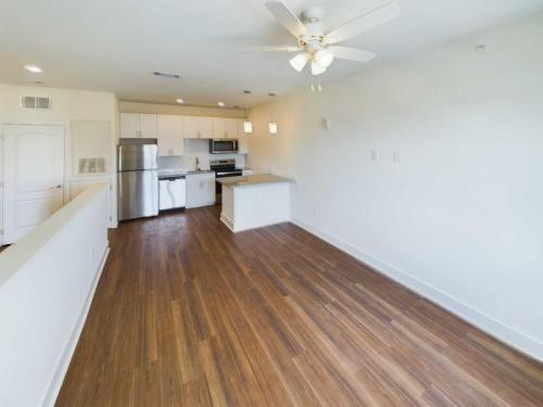 Apartments in Lebanon, IN A bright, empty kitchen with white cabinets, a stainless steel refrigerator, oven, and microwave, and a small island. The kitchen opens to a spacious living area with wood floors and a ceiling fan—a perfect example of the modern amenities found in apartments for rent in Lebanon, Indiana.