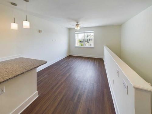 Apartments in Lebanon, IN A bright, empty room with wooden flooring, a ceiling fan, a window, and a kitchen counter with two pendant lights awaits you. White walls and a half-wall railing separate the kitchen and living area. Discover more such inviting spaces among the apartments for rent in Lebanon, Indiana.